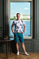 Tall handsome man wearing white t-shirt and blue shorts standing by big window in interior