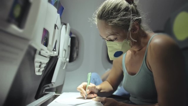 Woman travel caucasian at plane with wearing protective medical mask. Girl tourist at aircraft with protect respirator, fill out a registration form about her health. Coronavirus sars-cov-2 covid-19.