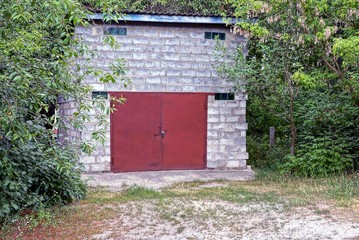 one white cinnamon garage with closed red iron gates among greenery and trees outside