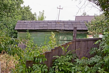 old rural barn and gray wooden fence overgrown with green vegetation and trees