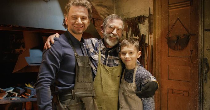 Portrait shot of happy joyful Caucasian old man with son and grandson hugging and smiling to camera in smithy worksop. Male family of forge craftsmen. Three generations of smiths.