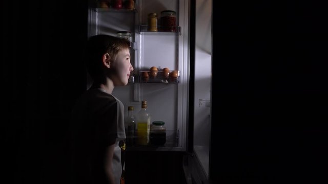 Shooting 4K . A young hungry boy eats at night . Hunger and gluttony from the refrigerator at night . A teenage boy looks into a full refrigerator at night .
