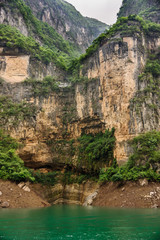 Wuchan, China - May 7, 2010: Dragon Gate Gorge on Daning River. Portrait of tall brown and black cliffs descending into emerald green water. Green foliage on top and bottom. Silver sky.