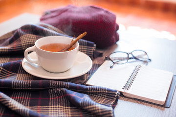 Close-up of a hot cinnamon tea, a notebook and glasses on a blue scarf over a table with a red sweater behind