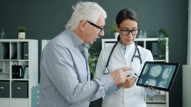 Young woman in uniform is showing patient MRI images on tablet screen talking holding gadget. People, healthcare and modern devices concept.