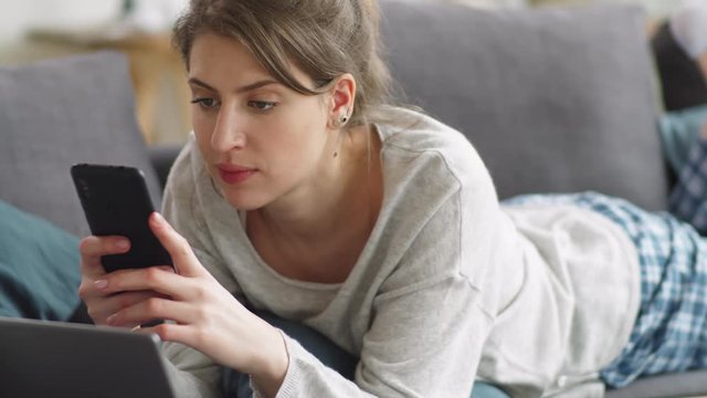 Tracking shot of young woman lying on stomach on couch in living room and surfing the Internet on smartphone while spending time at home during quarantine