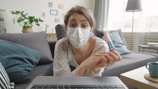Young woman in protective medical mask lying on couch in living room, looking and waving at camera, speaking and rubbing hands with sanitizer while video calling on laptop during quarantine
