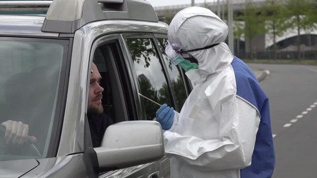 Health care worker in protective hazmat suit (and gloves, mask, and glasses) takes sample from coronavirus Covid-19 patient, through window of an SUV in a drive thru test clinic
