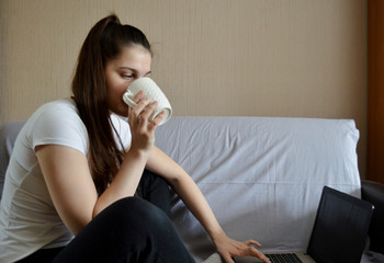 Young beautiful girl with dark hair sits with a laptop and coffee. Work from home. Remote work....