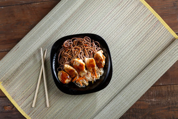 Black plate with soba noodles with chicken and vegetables in teriyaki sauce and chopsticks on a bamboo mat.