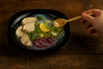 Black plate of Vietnamese Pho soup with chicken on a wooden table with a spoon.