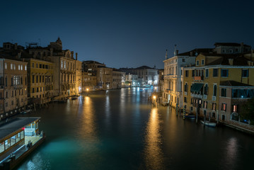 A scenic view of a beautiful Venetian canal at night with colourful architecture, a bridgeand reflections of boats running along the water in the town of Venice, Italy