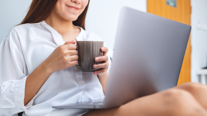 Closeup image of a beautiful woman using and working on laptop computer , drinking coffee while sitting on a white cozy bed at home