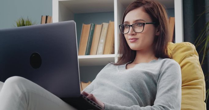 Charming young woman in eyeglasses and casual clothing sitting comfortably on beanbag chair and using personal laptop. Concept of freelance and remote work