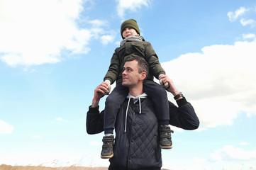 son in a knitted hat and jacket in khaki sits on the neck of a young father in a black jacket against a clear blue sky and white fluffy clouds. Games with children. Happy fatherhood.