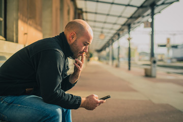 Side view of young bald male sitting on a bench and using a telephone and smoking while waiting for...