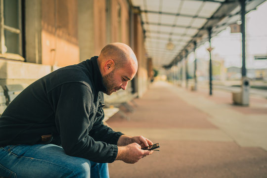 Side view of young bald male sitting on a bench and using a telephone and smoking while waiting for a train on an older style vintage train station.
