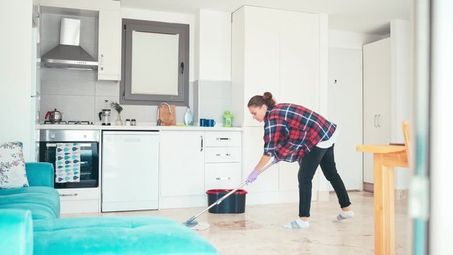 A cheerful young woman cleaning the kitchen mopping the floor doing housework in an apartment in protective gloves.