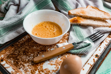 Whipped with a fork and mixed raw egg broken into a white bowl with yolk and protein, next to the shell, fork and towel on a rusty metal background as at home in the village, everything simple and ord