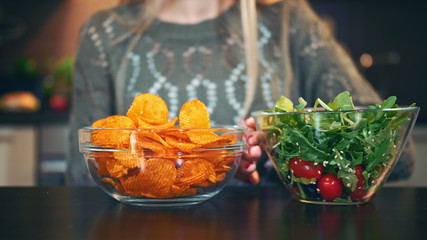Close-up shot of lady preferring crisp to salad. Attractive young woman choosing to eat healthy crisp for lunch while sitting at table in stylish kitchen and she eats these chips.