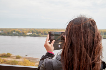 Fototapeta na wymiar Young woman with dark hair in black leather jacket taking selfies by cell phone on the river Bank in the autumn, spring or summer evening