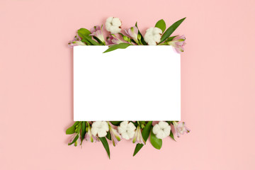 Paper card mockup with frame made of green leaves, cotton, alstroemeria flowers on a pink pastel background. Holiday floral concept.