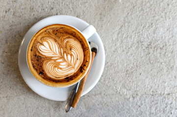 Hot cappuccino coffee in white ceramic cup with cinnamon and spoon on saucer, placed on concrete table in cafe