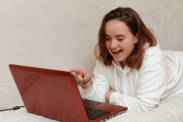 A young girl lies at home on the sofa with a computer and communicates via Skype with friends,...