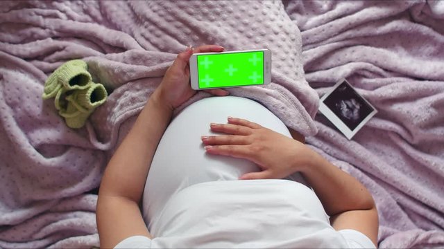 A pregnant woman is stroking her stomach and holding a phone with a chromakey in one hand. Top view of a pregnant woman's belly stroking her belly and holding an ultrasound photo