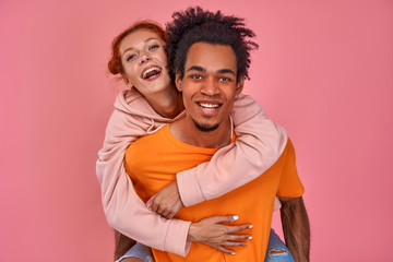 Naklejka premium Photo of playful African American man gives piggyback ride to red-haired girlfriend, have fun together, smile happily, pose over pink background. Happy man carries a woman on the back