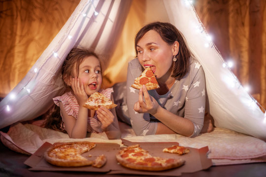 Family bedtime. Mom and daughter eat pizza in a tent. Pretty young mother and lovely girl having fun in children room.