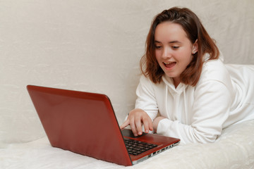 A young girl lies at home on the sofa with a computer and communicates via Skype with friends,...