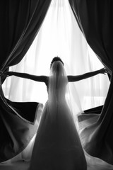 Bride with bridal veil standing in front of the window and opening curtains.  Black ad white photo