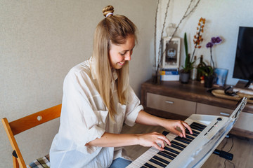 Woman is quarantined at home coronavirus. Attractive girl in a white shirt and jeans staying at home on self-isolation and playing the electronic piano - synthesizer. Hobbies - what to do at home?
