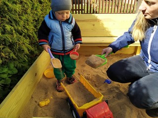 little boy with mom playing in the sandbox surrounded by greenery with toy cars and shovels on a sunny day