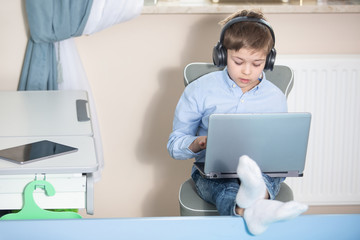 work from home - smiling young boy in blue shirt sits at his desk working on his notebook computer wearing headphones