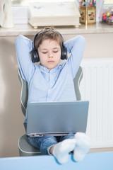 work from home - smiling young boy in blue shirt sits at his desk looking at his notebook computer wearing headphones