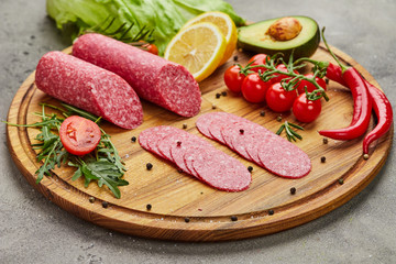 Traditional thinly sliced salami on a wooden cuttingboard  with spices, lettuce, avocado and tomatoes. Cold meat plate