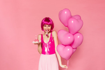 Image of woman blowing in party horn while holding balloons and cake