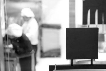 Primo piano cartello nero su cui poter scrivere e sullo sfondo sfocati una cucina con dei cuochi a...