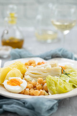 boiled cod fish with boiled vegetables and egg on white dish and glass of wine on ceramic background