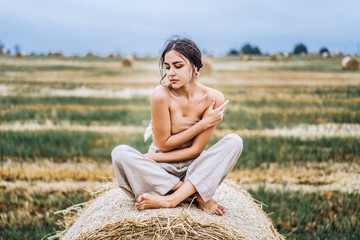 Naklejka na ściany i meble Brunette in linen pants and bare shoulders sitting on a hay bales in warm autumn day. Woman looking at camera. Behind her is a wheat field