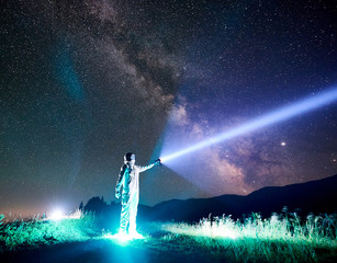 Full length of astronaut illuminating beautiful starry sky with flashlight. Cosmonaut wearing white space suit and helmet while standing on glowing grass. Concept of light, galaxy and astronautics.