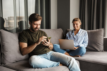 Photo of joyful couple using laptop and mobile phone while sitting