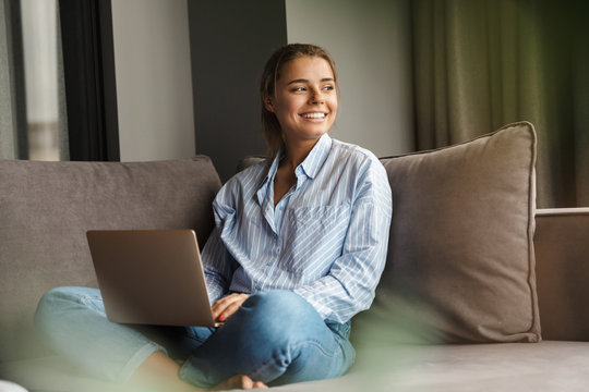 Image of joyful woman using laptop and smiling while sitting on sofa