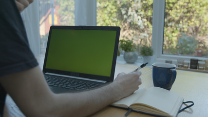 Working from home using a green screened laptop on the kitchen table with pens and a pad