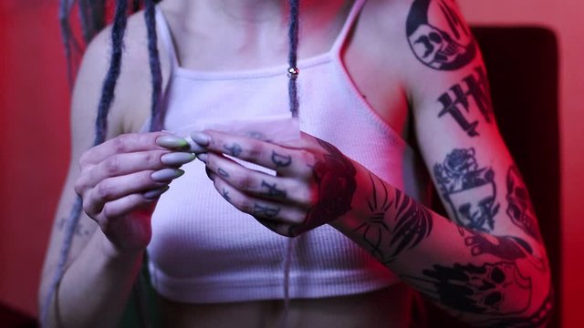 Attractive young girl wraps cannabis in a piece of paper for smoking. freak with dreadlocks and tattoos close-up in a neon light