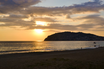 Golden sunset over the Mediterranean Sea. The setting sun shines through the clouds, and a golden path is visible above the ocean. Silhouetted mountains on the horizon.