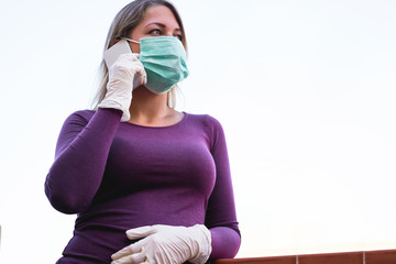 Young woman talking with mobile phone outdoor wearing face mask and gloves for Covid-19 virus protection - Girl speaking with smartphone outside during quarantine isolation - Focus on left hand glove