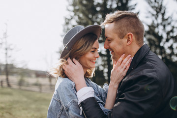 Country wedding. A guy in a leather jacket and a young girl in a gray-blue dress and hat hug and kiss on a background of sky and mountains at sunset